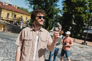 Wall Mural - tour guide in sunglasses and headset smiling and pointing with finger near blurred multiethnic touri