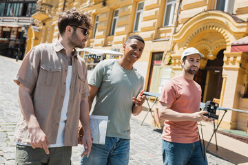Wall Mural - joyful interracial travelers with city map and vintage camera walking with tour guide in sunglasses 