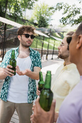 Wall Mural - young and trendy man in sunglasses pointing at beer bottle near blurred interracial friends in city 