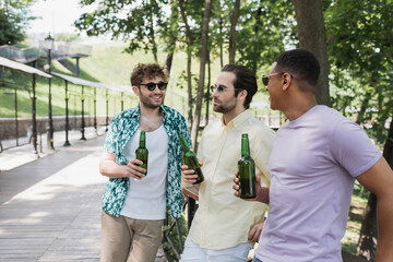 Wall Mural - carefree interracial friends in stylish summer outfit holding beer bottles and talking in urban park