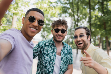 Wall Mural - Positive interracial friends in sunglasses pointing at camera in summer park.