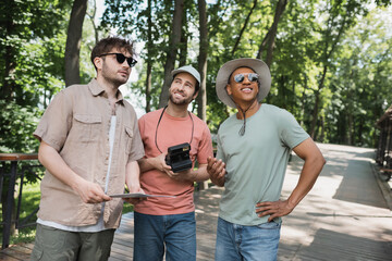 Wall Mural - cheerful bearded tourist with vintage camera looking away near multicultural men in green city park.