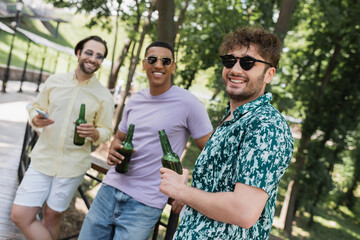 Wall Mural - positive man holding beer bottle near interracial friends in sunglasses in summer park.