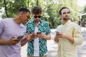 Wall Mural - joyful african american man pointing with finger near trendy friends using cellphones in summer park
