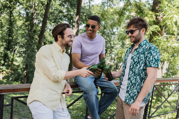 Wall Mural - cheerful multiethnic friends in sunglasses clinking beer in summer park.