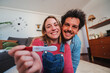 © Jose Calsina - Happy caucasian young couple smiling excited showing at camera their positive pregnancy test at home. Husband and wife celebrating that they are expecting a baby holding a predictor in their hands