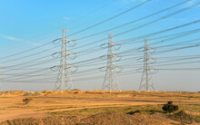 Large Steel Electricity Power Pylons With Cables, Blue Sky Background, Afternoon Sun Shines On Rather Desert Landscape Around