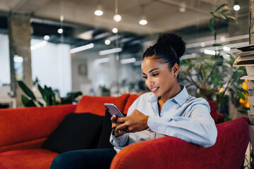 Wall Mural - African businesswoman sitting on a couch, using a mobile phone.