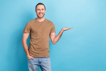 Poster - Photo of young smiling business man wear brown t-shirt promoter hold palm demonstrating new product sale advert isolated on blue color background