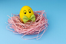 A Yellow Easter Egg With Drawn Eyes Wearing A Green Bow Tie Dyed With Turmeric Lies In A Pink Nest On A Blue Background.