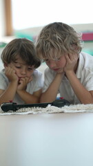 Canvas Print - Two small boys watching media on screen laid on floor with hands on chin. Children consuming content online close up faces in Vertical Video