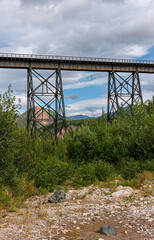 Denali Park, Alaska, USA - July 24, 2011: Portrait. Tall slender railway bridge over Nenana River in its rocky bed with green forests on both sides under blue cloudscape