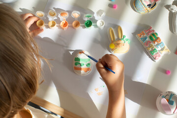 Wall Mural - A cute little girl is sitting at a table and doing Easter crafts, coloring Easter figurines. Creativity and hobbies.