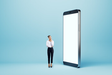 Poster - Thoughtful young businesswoman looking at big empty white smartphone on blue background. Technology, innovation and communication concept. Mock up.