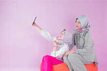 Two Teenage Women In Hijabs Taking Selfie With Cell Phones Sitting On Orange Bean Chairs, Isolated Pink Indoor Studio. Negative Space