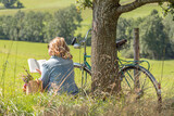 Fototapeta Sawanna - Relaxed blonde Woman reading book under tree. Retro bicycle and basket with flowers.