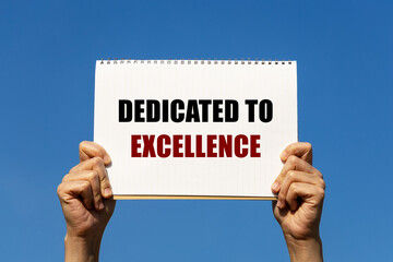 Dedicated to excellence text on notebook paper held by 2 hands with isolated blue sky background. This message can be used as business concept about dedicated to excellence.