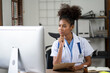 © Parichat - Portrait of female African American doctor sitting in her office at clinic. working with dewsktop computer.