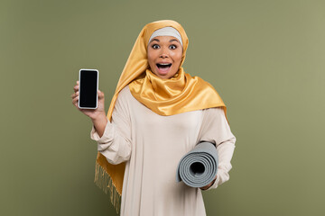 Excited multiracial woman in hijab holding smartphone and fitness mat on green background.