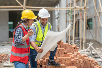 Asian female civil engineer and caucasian male architect wears safety vest with helmet discuss and look at blueprints in housing estate construction site