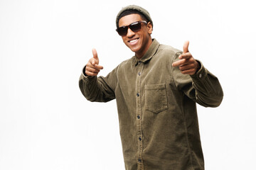 Playful african-american guy in sunglasses, trendy hat and shirt pointing at camera isolated on white, positive cheerful man enjoying having fun, choosing you, pretending making shot from imagine guns