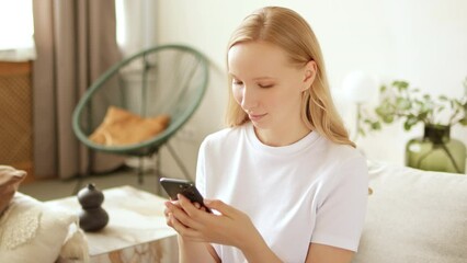 Young blonde woman is using phone swiping social media and smiling at home. Texting online with friends through a messenger or plays mobile games app.