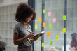© SOMKID - Young African American stylish woman working on project plan using sticky papers notes on glass wall, people meeting to share idea, positive thinking workshop and business design planning concepts.