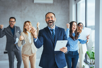 Wall Mural - Successful business group celebrating an achievement at the office with arms up and looking at the camera smiling. Shot of a group of young businesspeople huddled together in solidarity