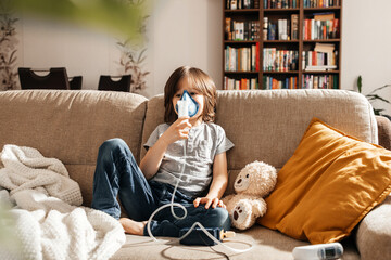 a little boy makes an inhalation at home sitting on the couch using a nebulizer and a mask. preventi