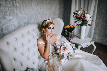 A beautiful young blonde bride in a white lace dress with a crown, diadem sits on a sofa in the studio, indoors with a bouquet of flowers in her hands. Wedding photography, portrait.