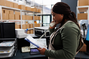 Wall Mural - African american worker talking at landline phone with remote customer discussing order details. Storehouse employee holding clipboard, analyzing goods inventory report in storage room.