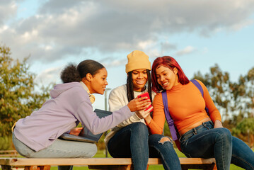 multiracial group of 3 female friends sitting on a bench on a university campus using a cell phone and a laptop. classmates having a good time during a break