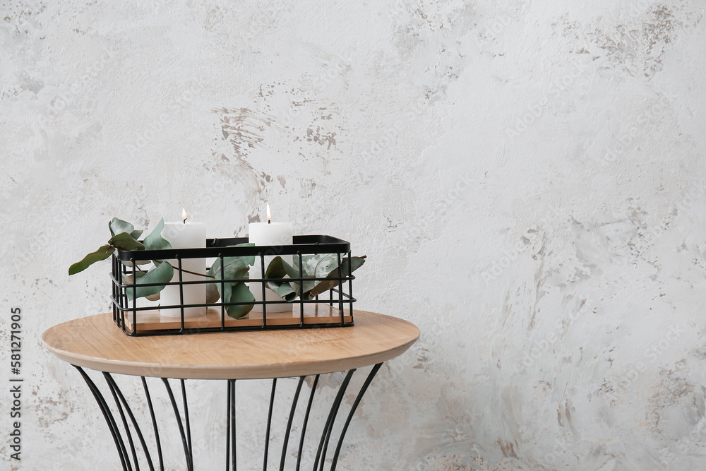 Basket with burning candles and eucalyptus branch on table near grey grunge wall