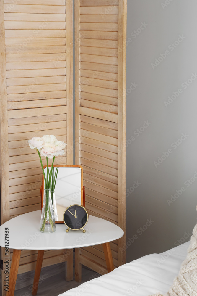 Vase with ranunculus flowers, alarm clock and mirror on table in bedroom
