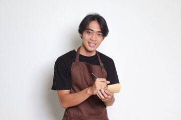 Portrait of attractive Asian barista man in brown apron taking order, writing on menu book list. Isolated image on white background