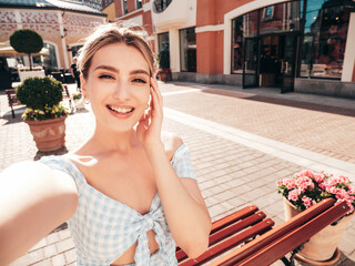 Wall Mural - Young beautiful smiling hipster woman in trendy summer dress clothes. Sexy carefree model posing on the street background at sunset. Positive blond female outdoors. Taking Pov selfie photos