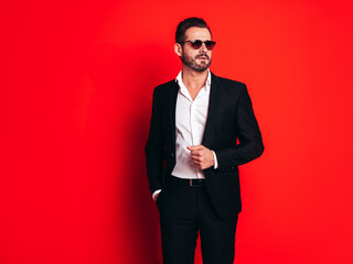 Wall Mural - Portrait of handsome confident stylish hipster lambersexual model. Sexy modern man dressed in black elegant suit. Fashion male posing in studio, isolated on red. In sunglasses