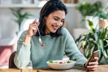 Beautiful young woman eating a bowl of salad while usingh her mobile phone in the living room at home.