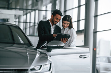 Showing information in the graphic tablet. Man in formal clothes is consulting woman about the automobile in the car dealership