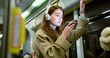© ihorvsn - Side-view portrait of attractive happy caucasian female using public transport standing holding handrail watching videos on smartphone and wireless headphones. Charming young woman travelling by bus.