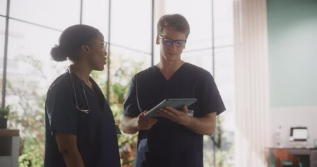 Wall Mural - Two Young Doctors Standing in a Medical Facility in the Hospital, Talking and Sharing Information on Tablet Computer. Modern Hospital with Diverse Multiethnic Professional Health Care Personnel