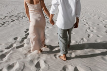 Walking Young Couple In Love. European Girl And Muslim Guy Hold Hands And Go Back To Camera, Climb Sand Hill Together And Stand Opposite Each Other, On Sandy Desert Hill Against Blue Sky And Sunset On