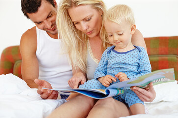 Wall Mural - We all enjoy storytime. Two young parents sitting on the bed reading a story book to their young son.