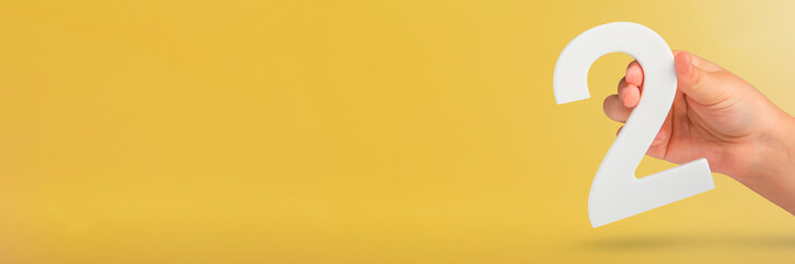 Fototapete - Number two in hand. Hand holding white number 2 on yellow background with copy space. Concept with number two. 2 percent, birthday 2 years old, couple, two, double