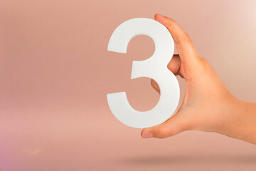Fototapete - Number three in hand. Hand holding white number 3 on red background with copy space. Concept with number three. 3 percent, birthday 3 years, third grade, triple