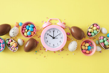 Wall Mural - Easter celebration concept. Top view composition of chocolate eggs сolorful candies in paper molds sprinkles on pastel yellow background with pink alarm clock in the middle