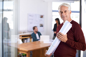 Poster - Design minds at work. Portrait of a mature male architect holding blueprints with his colleagues working in the background.