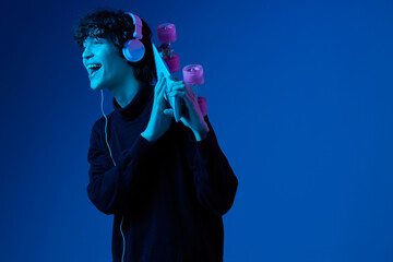 Wall Mural - Teenage man wearing headphones listening music and dancing and singing with skateboard in hand over his head, hipster lifestyle, blue background, neon light, style and trends, mixed light, copy space
