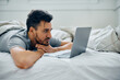 Smiling man using laptop while relaxing in bedroom.