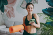 © ViDi Studio - Young strong athletic fitness trainer instructor woman wearing green tracksuit hold in hand yoga mat use mobile cell phone training do exercises at home gym indoor. Workout sport motivation concept.
